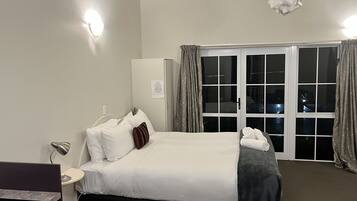 A hotel room with a bed, a nightstand, a desk with a lamp, a window with curtains, and a view of the outside.