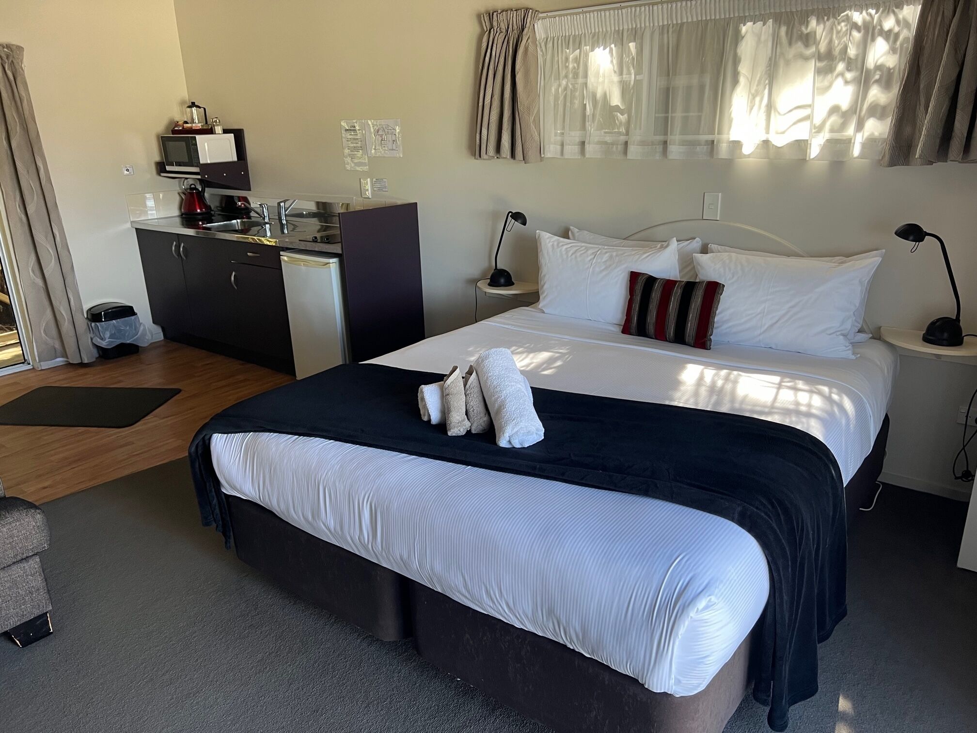 A hotel room with a bed, bedside tables, a desk with a microwave, and a small kitchenette.