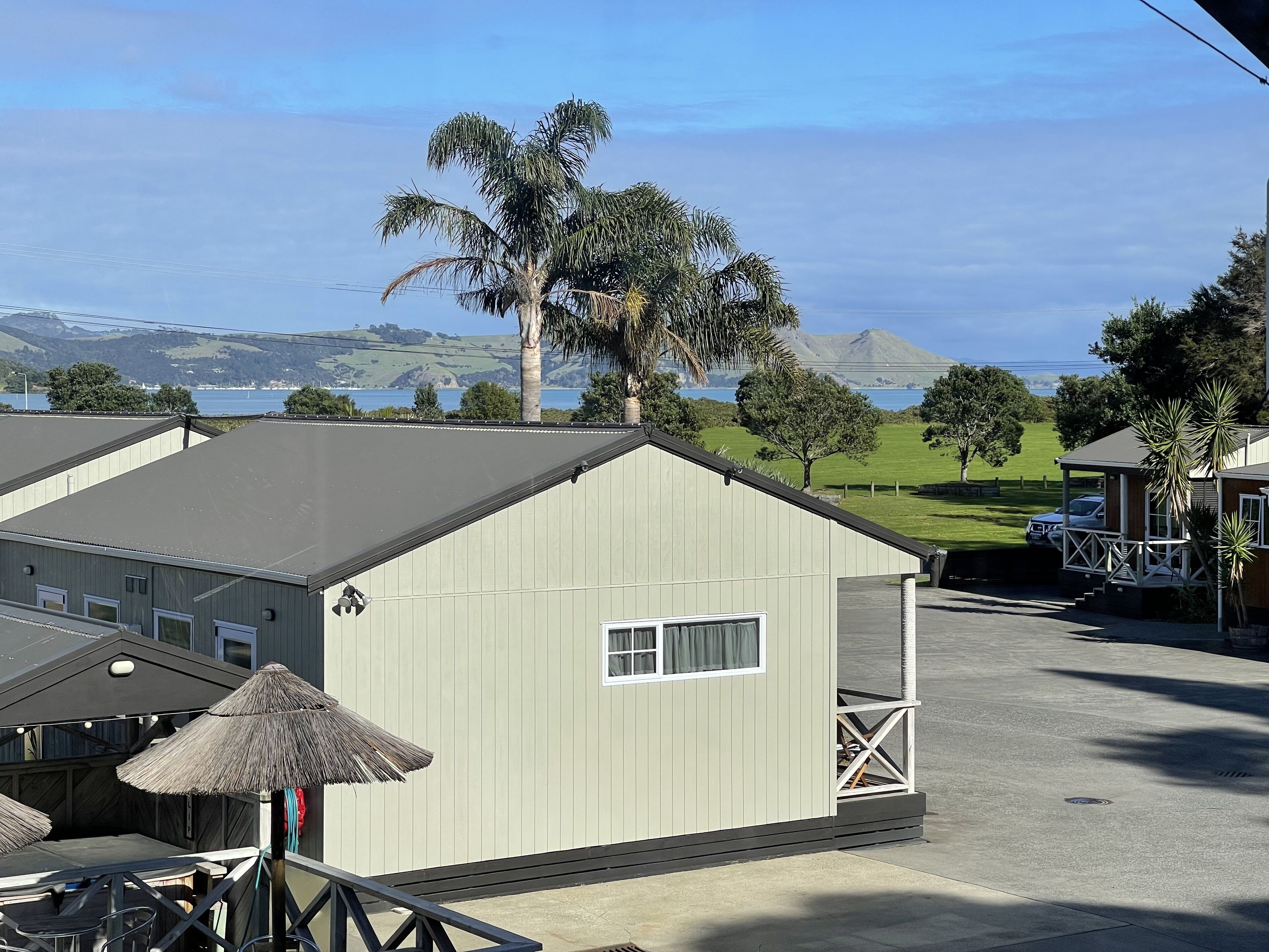 A small, single-story building with a balcony, a thatched umbrella, and a view of the ocean and hills in the background.