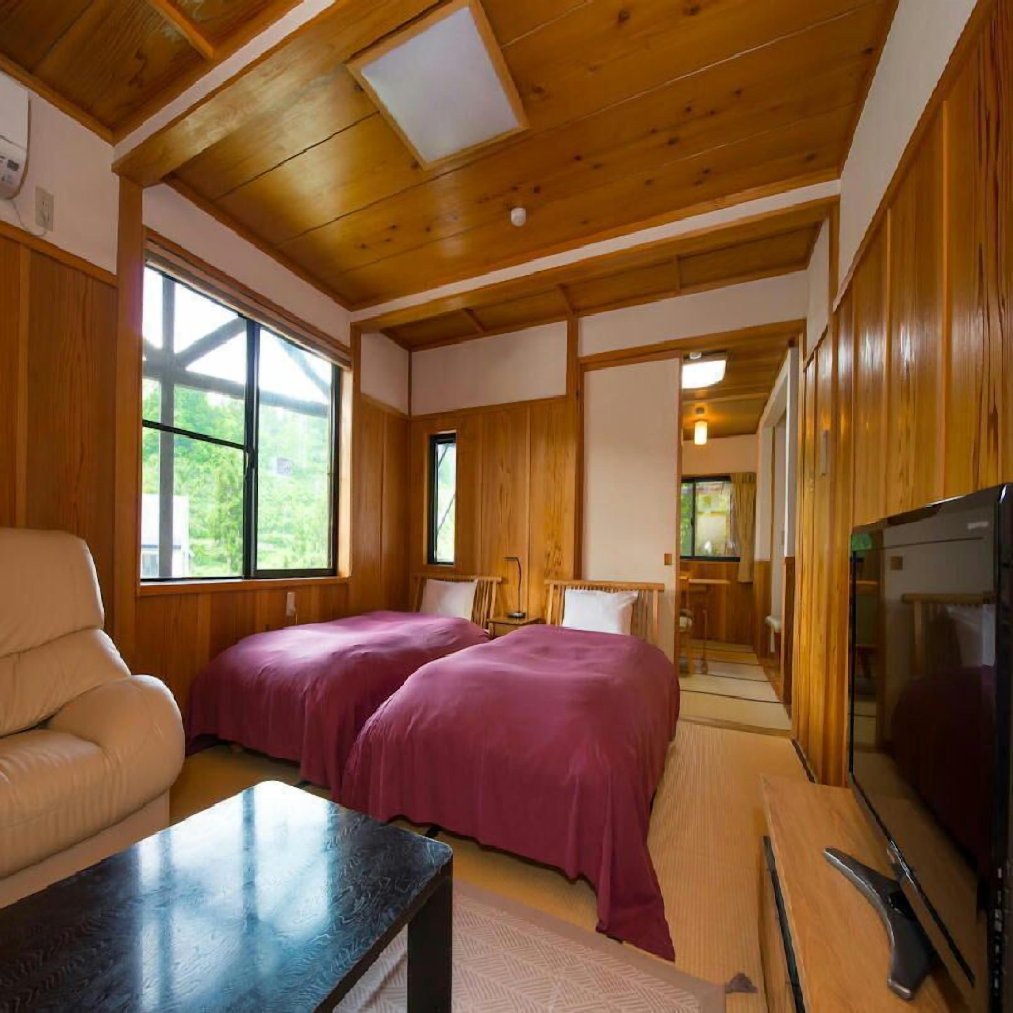 A cabin with wooden walls and ceiling, a bed with purple bedding, a chair, a table, and a TV.