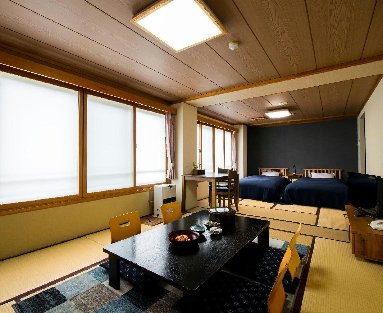 A room with a dining table, chairs, a TV, two beds, and large windows with curtains.