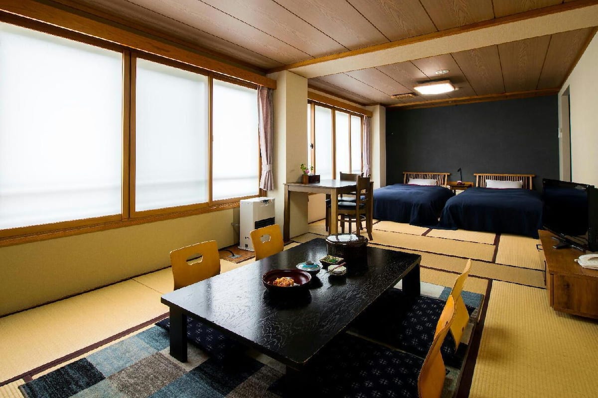A room with a dining table, chairs, a TV, two beds, and large windows.