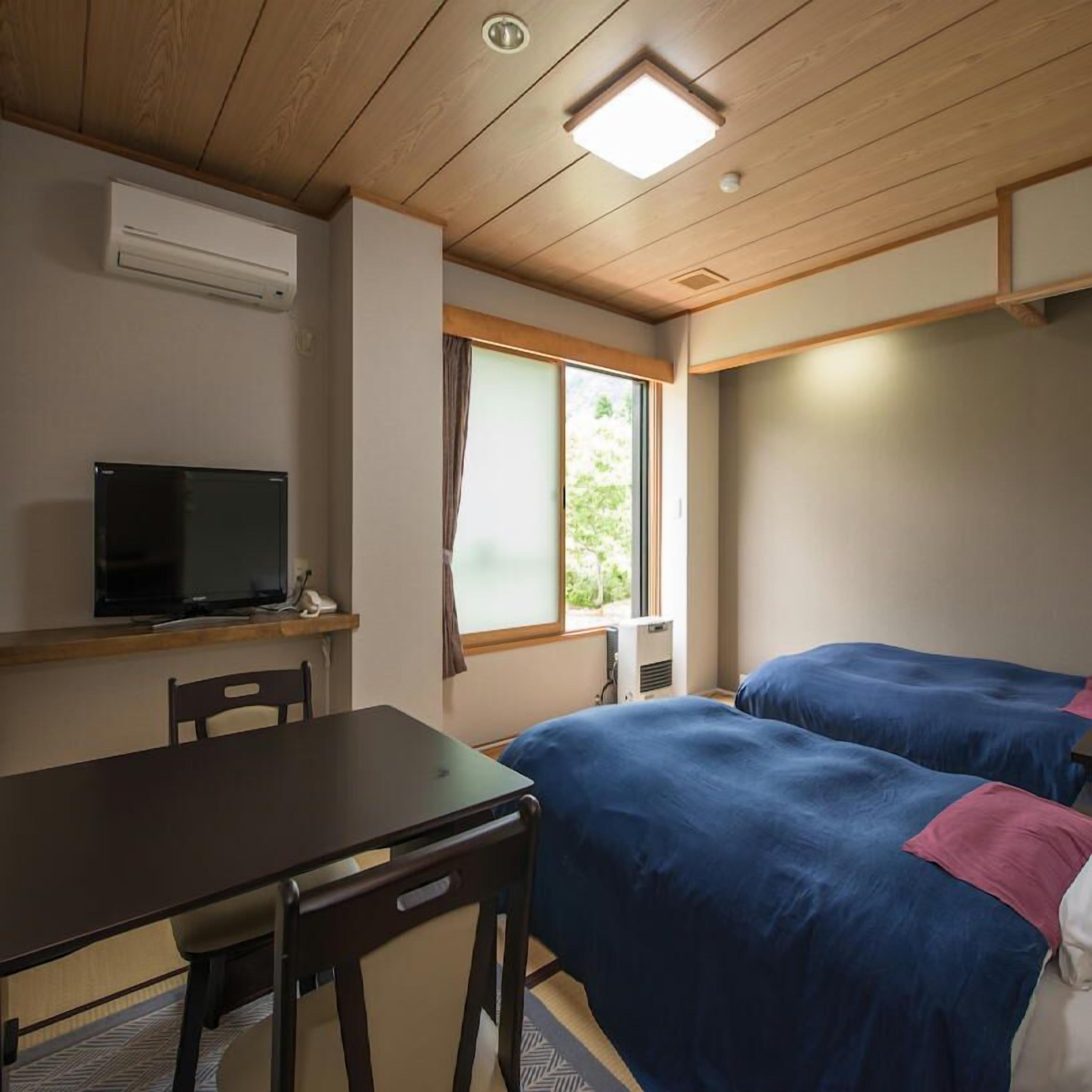 A room with a wooden ceiling, a TV on a shelf, a desk, two beds with blue bedding, and a window with curtains.