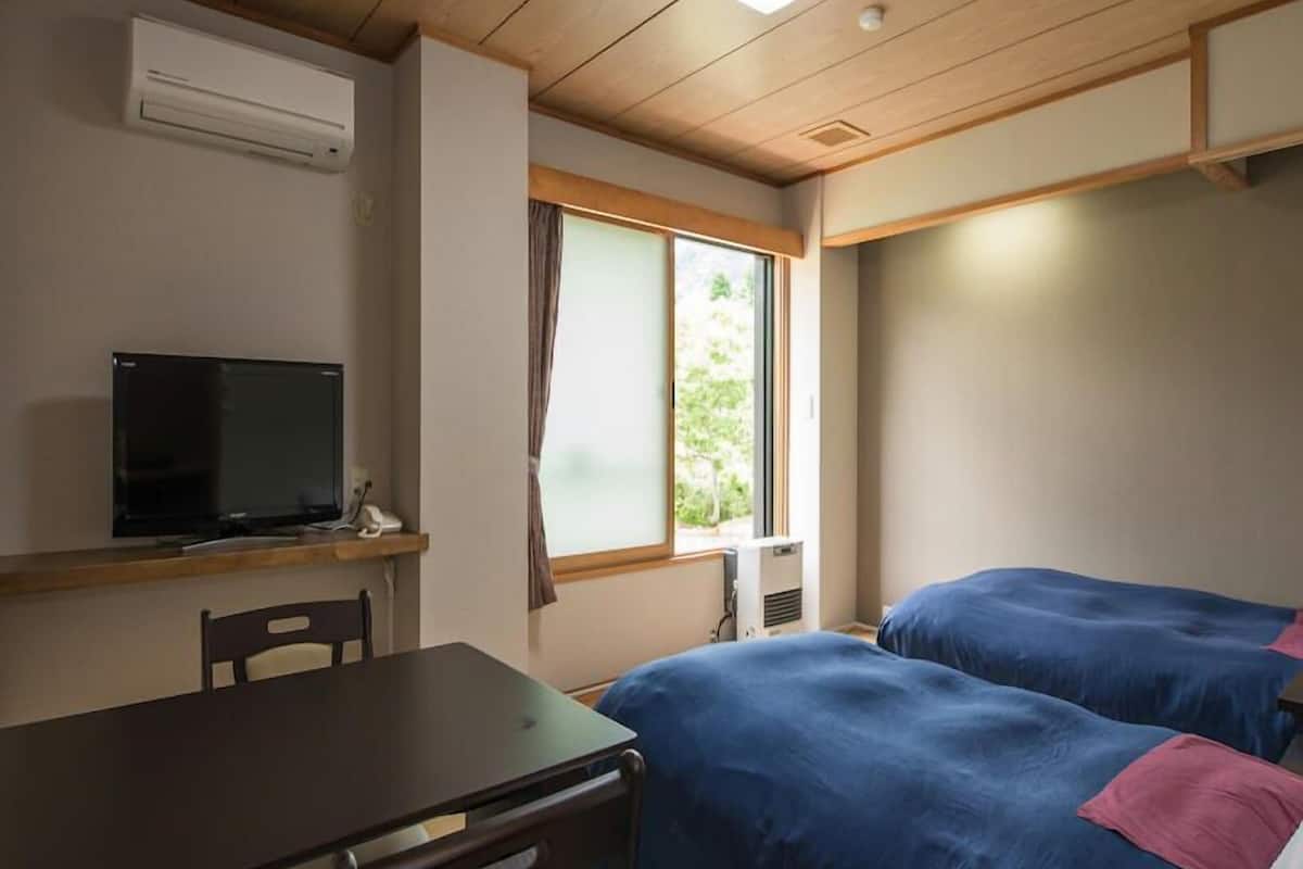 A room with a wooden ceiling, a TV on a stand, a desk, two beds with blue bedding, and a window with curtains.