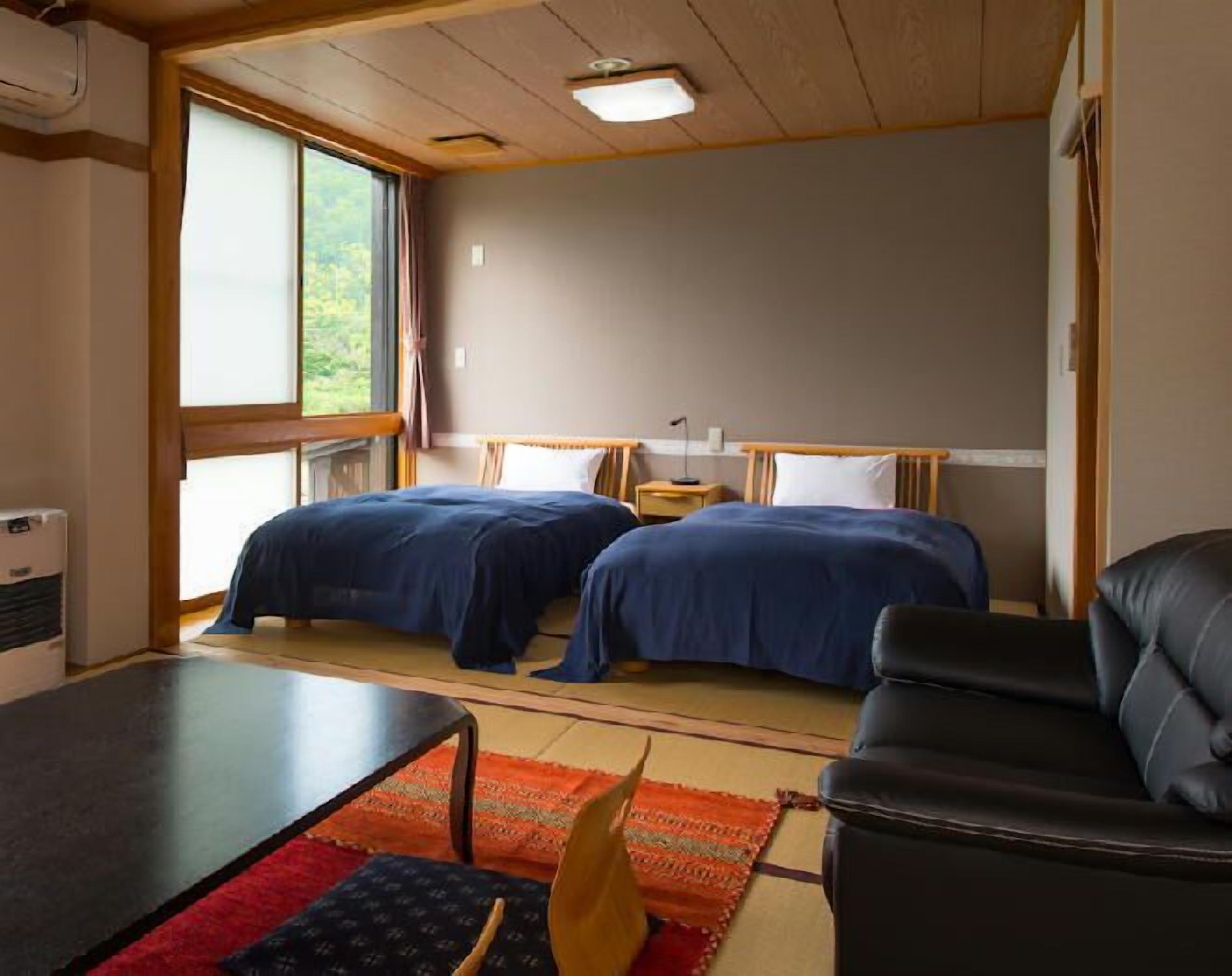 A room with two beds, a small table, a couch, and a window with a view of greenery.
