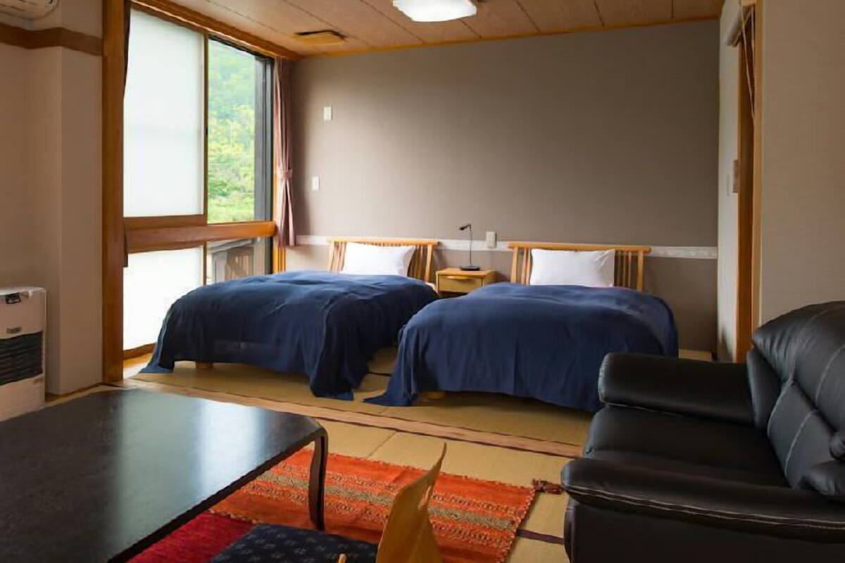 A room with two beds, a small table, a couch, and a window with a view of greenery.
