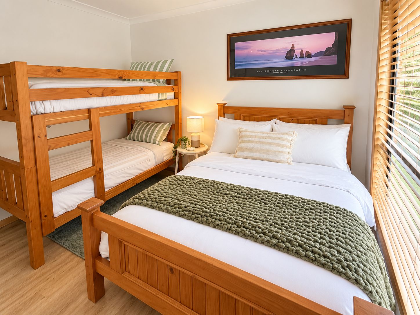 A room with a bunk bed, a single bed, a framed picture on the wall, and a window with blinds.