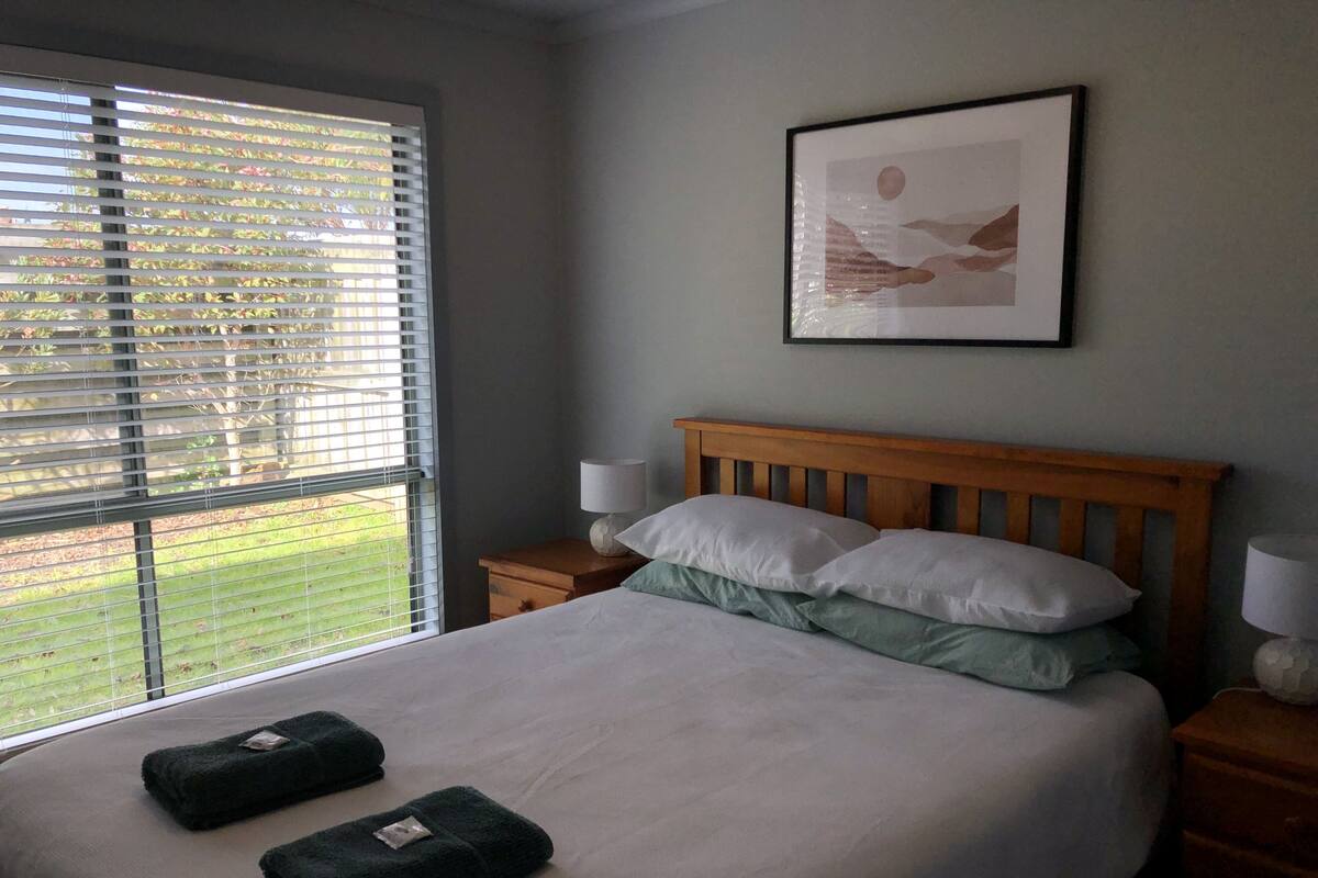 A neatly made bed with two pillows and two folded towels, a wooden headboard, a bedside table with a lamp, and a framed picture on the wall.