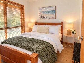 A bedroom with a wooden bed, two bedside lamps, a painting above the bed, and a window with blinds. - Seahorse Coastal Villas (Peterborough)