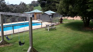 A backyard with a wooden swing set, a picnic table, a swimming pool, and a fenced area with a shed. - Neerim Country Cottages (Neerim South)