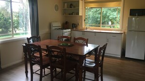 A dining area with a wooden table and chairs, a kitchen with a stove, sink, and refrigerator, and a window with a view of trees. - Neerim Country Cottages (Neerim South)