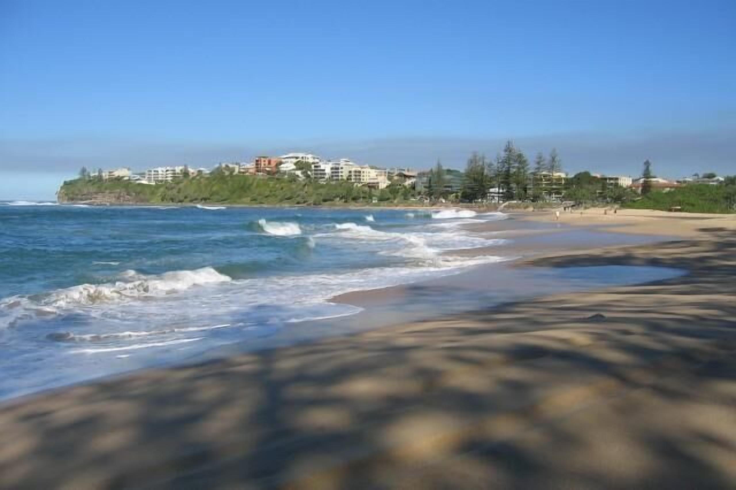 Une plage avec des vagues, un ciel dégagé et des bâtiments en arrière-plan.