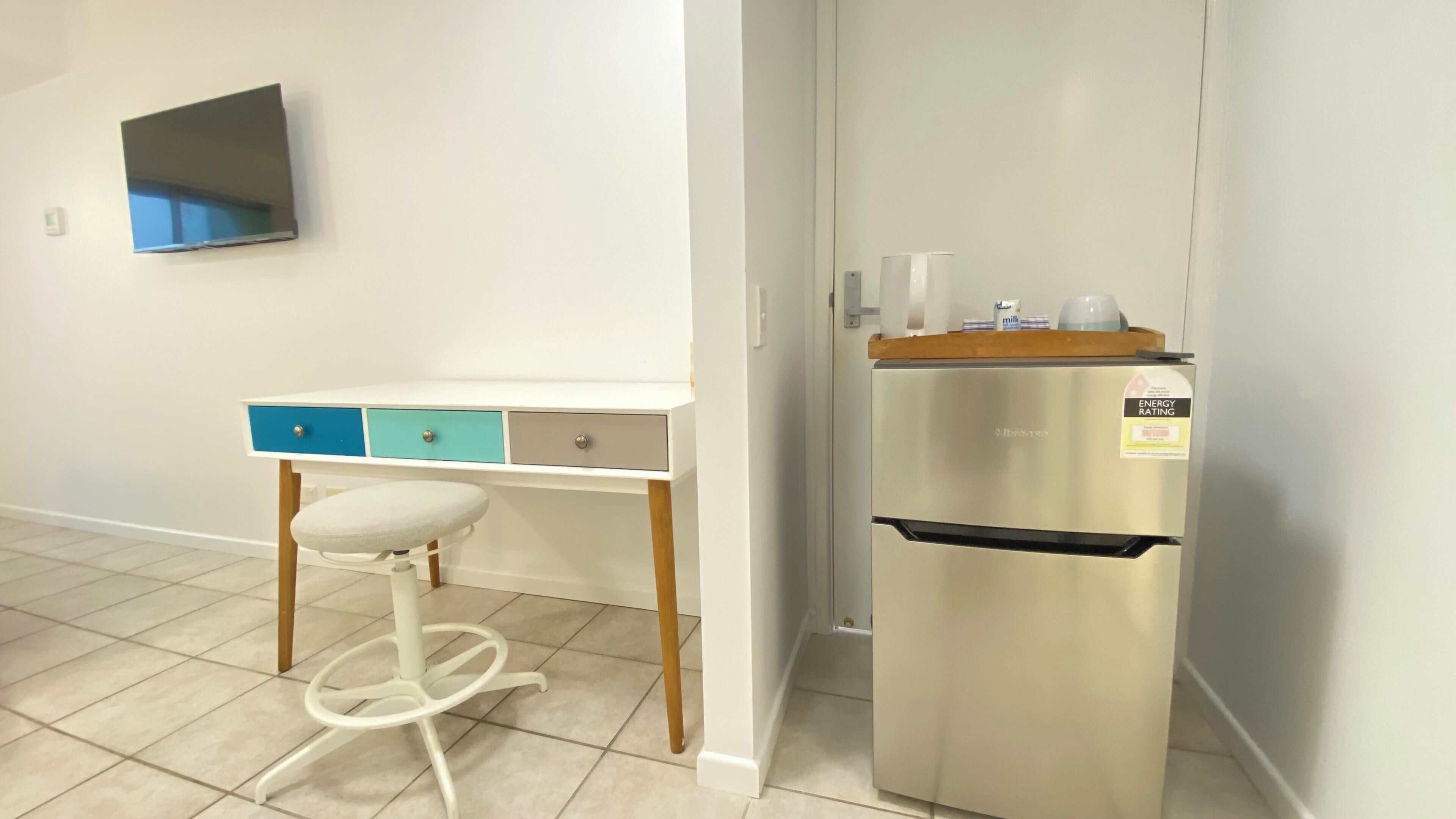 Una kitchenette con un refrigerador blanco, una mesita pequeña con cajones color turquesa y un taburete blanco.