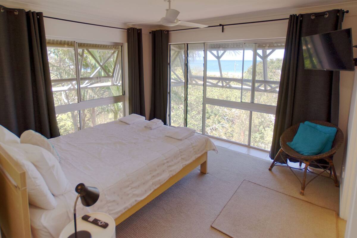 A bedroom with a bed, a chair, a TV, and large windows with a view of the beach.