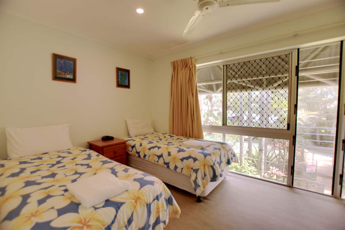 A hotel room with two beds, a window with a view of greenery, a ceiling fan, and a small table with a lamp.