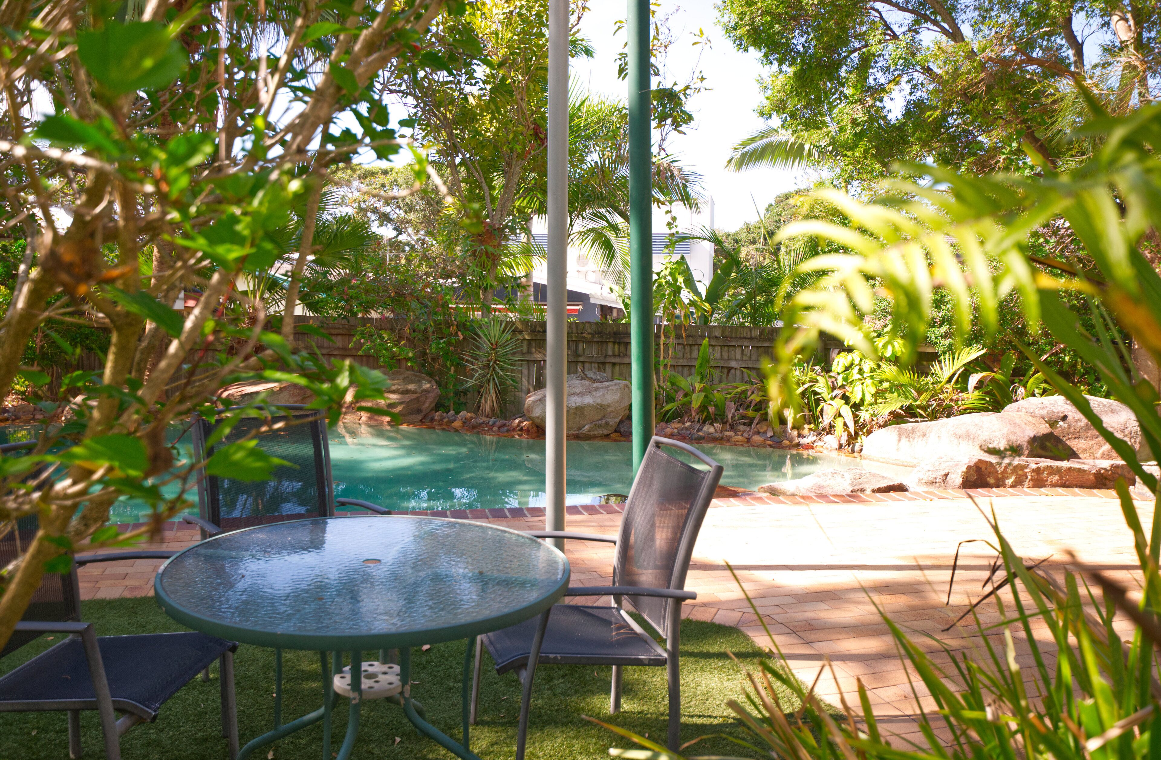 A glass-top table with metal chairs in a garden area with a pool.
