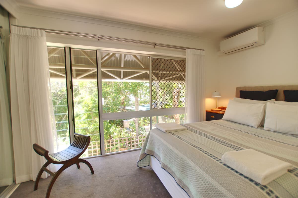A bedroom with a large bed, a bench, and a view through a sliding glass door.