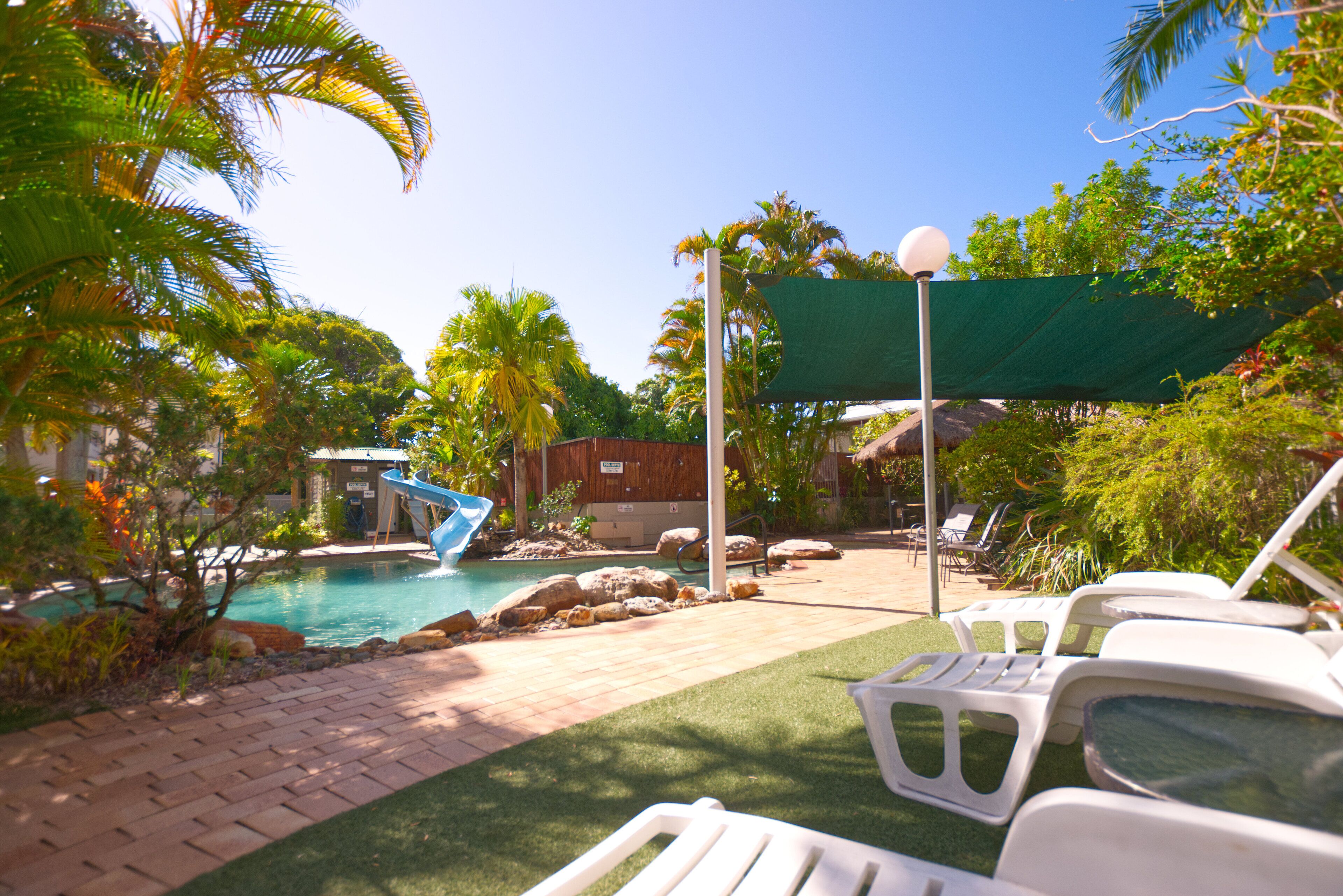 Zona de piscina con sillones de descanso, tobogán de agua y palmeras.