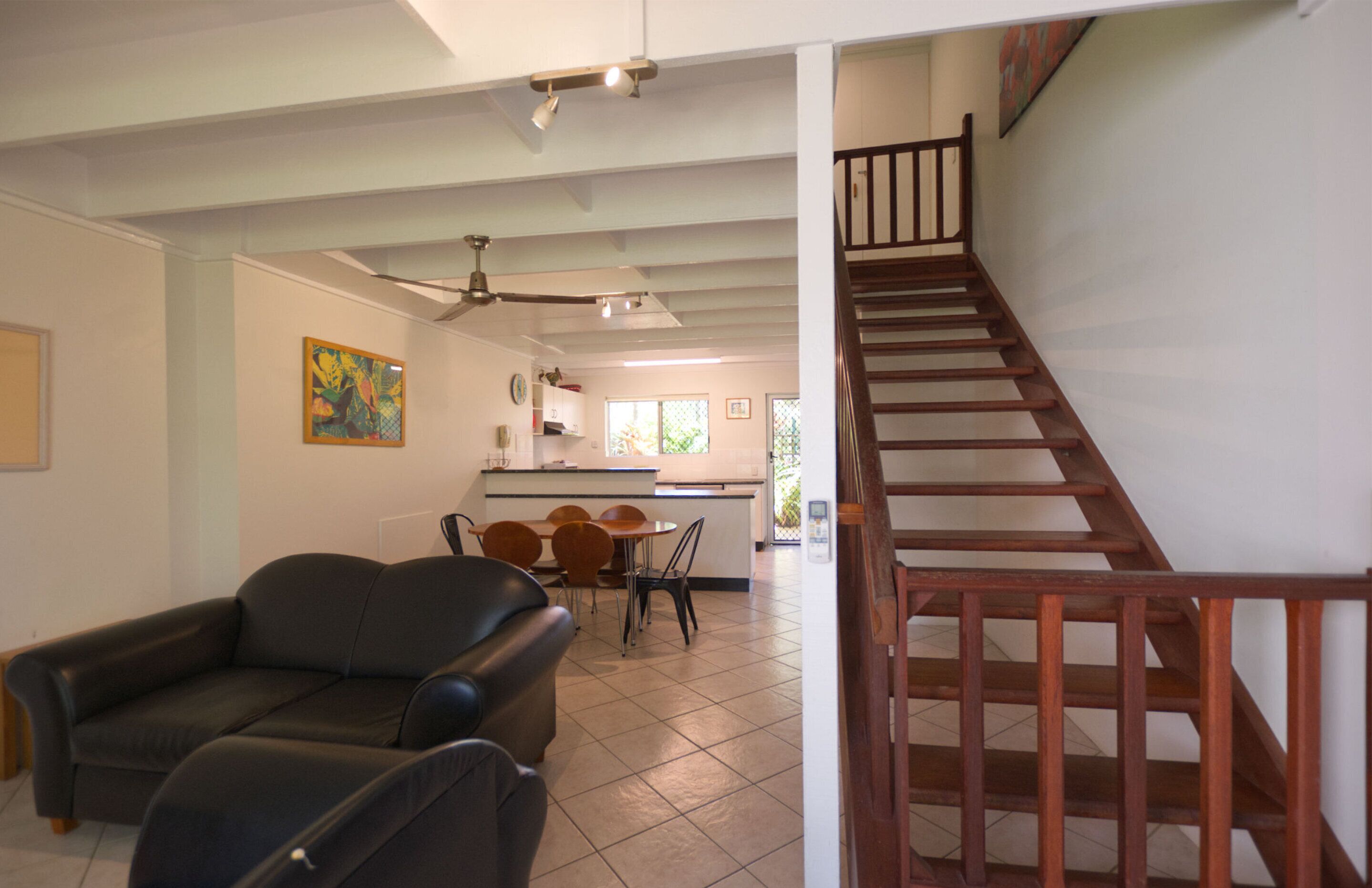 Un salon avec un canapé en cuir noir, un coin repas avec une table et des chaises, une cuisine à l’arrière-plan et un escalier en bois.