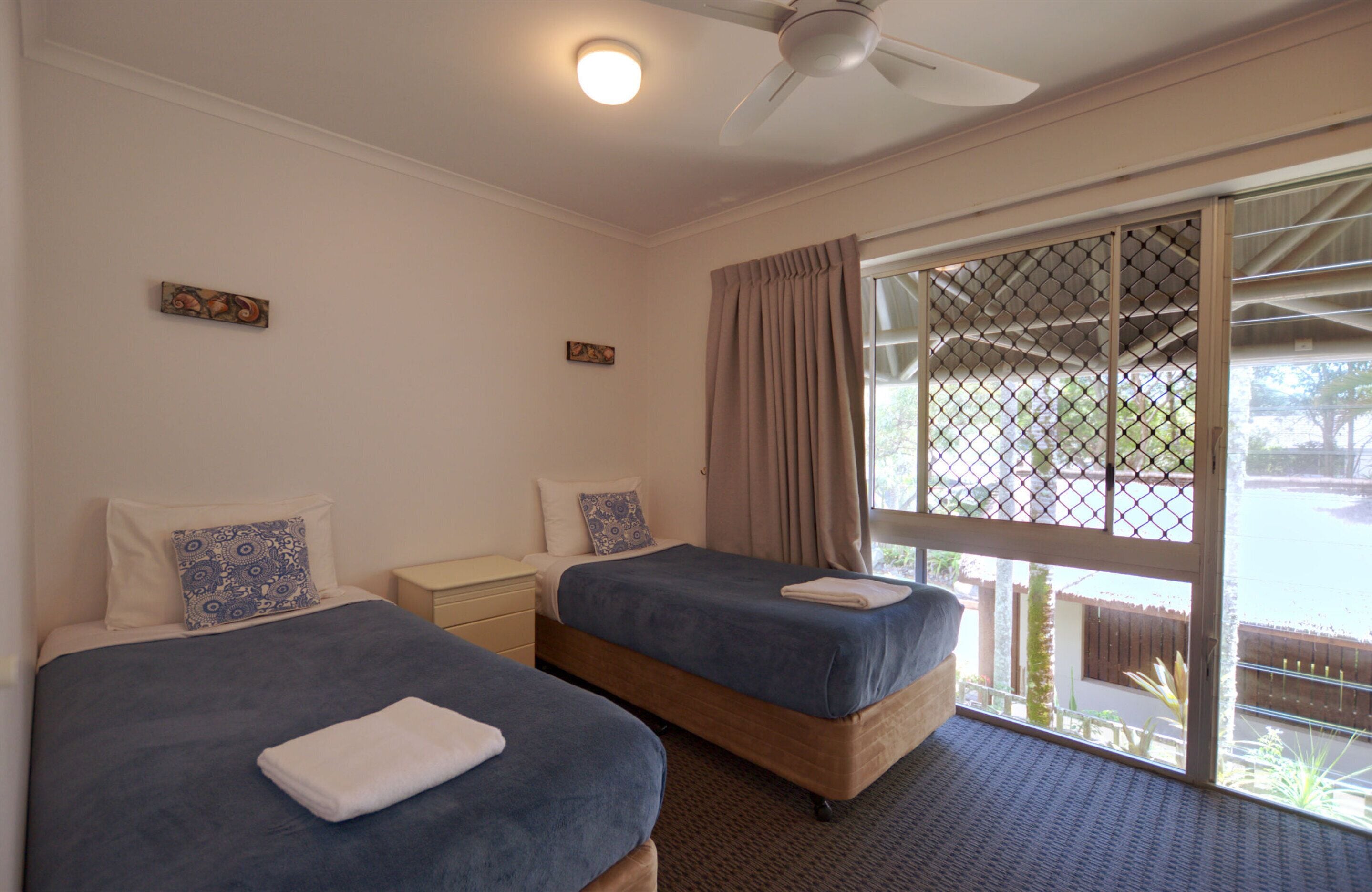A hotel room with two beds, a ceiling fan, and a large window with a view of the outdoors.
