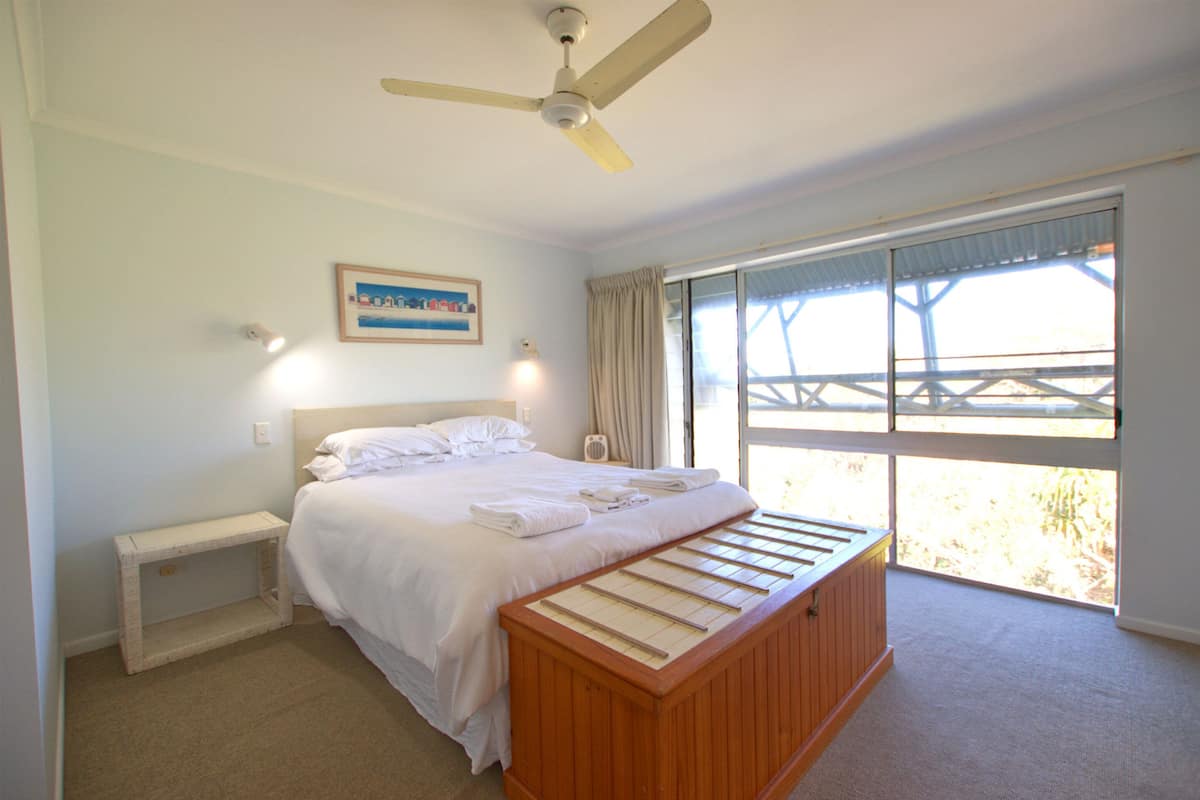 A bedroom with a bed, a ceiling fan, a bedside table, a window with curtains, and a painting on the wall.