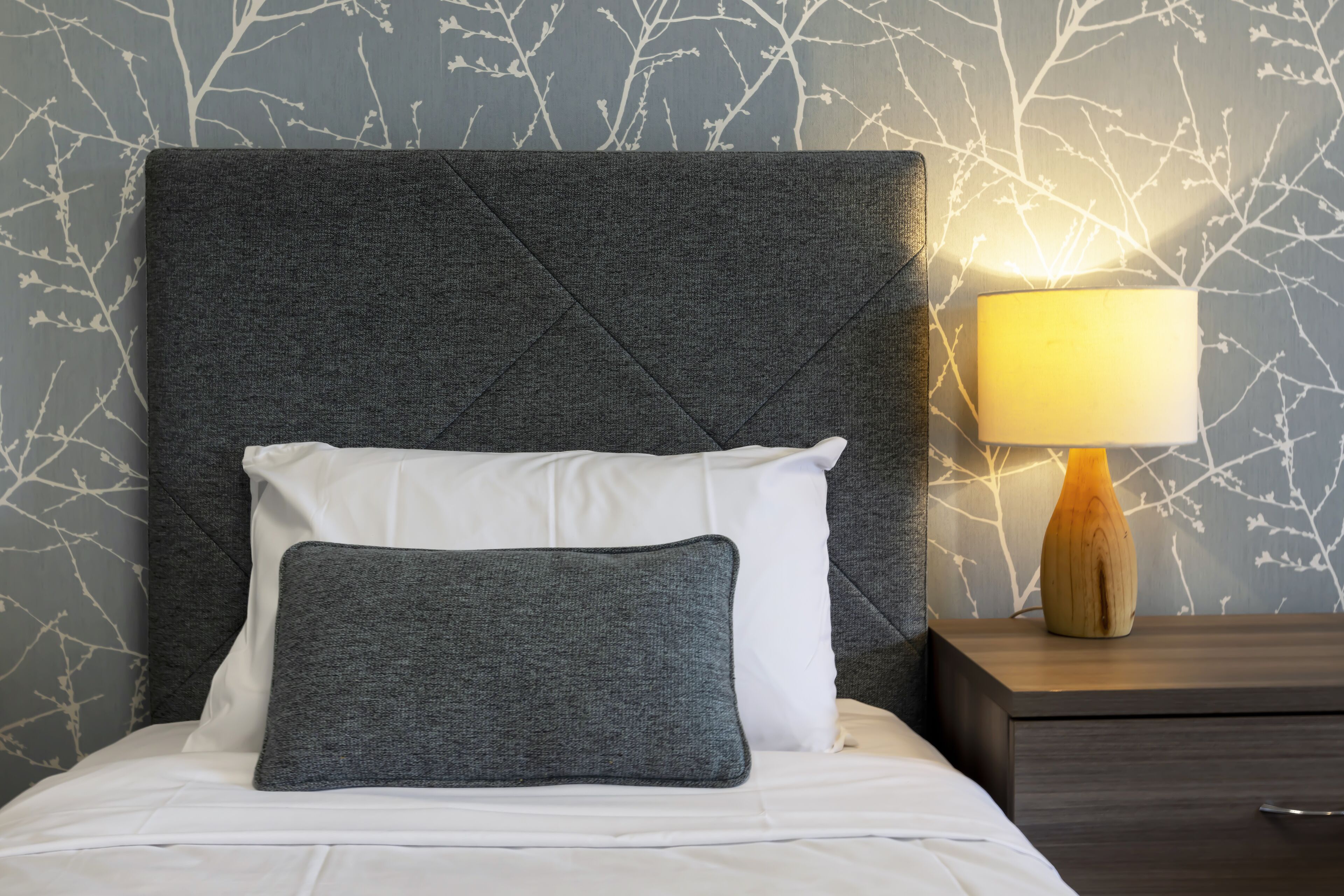 A neatly made bed with a dark headboard, a white pillow, and a grey pillow. A wooden bedside table with a lamp is to the right.