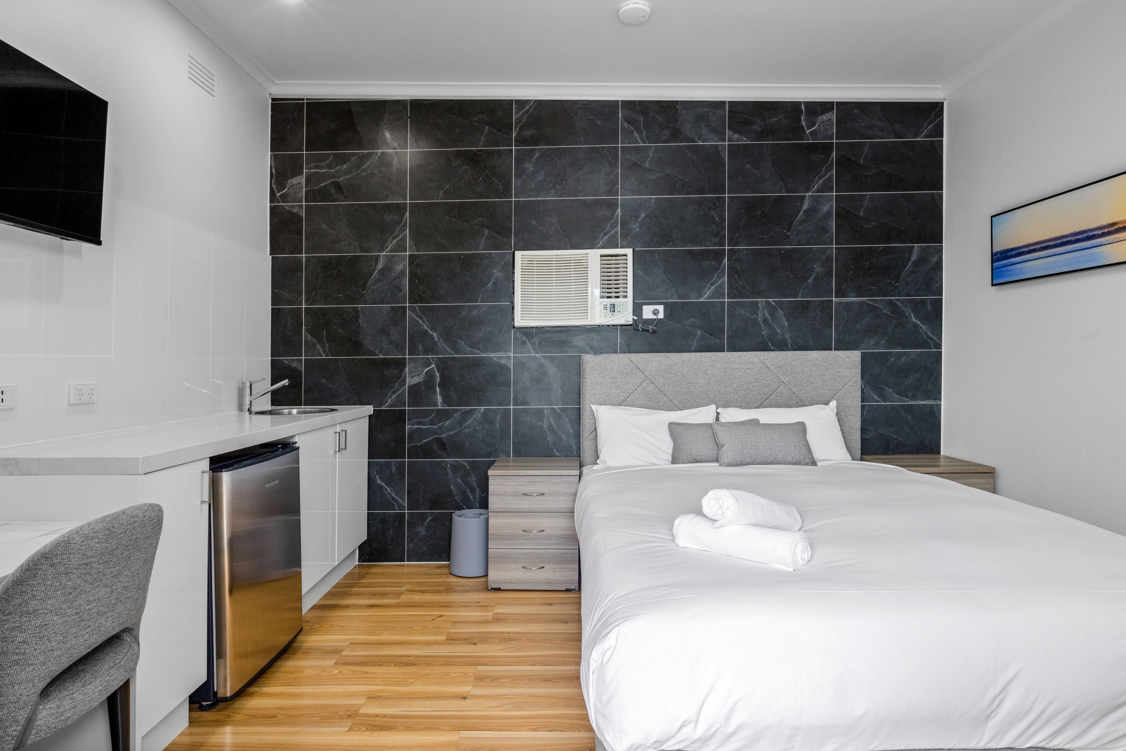 Una camera d'hotel moderna con parete rivestita in marmo, un letto con lenzuola bianche, una poltrona in grigio, un condizionatore d'aria integrato e un comodino in legno.