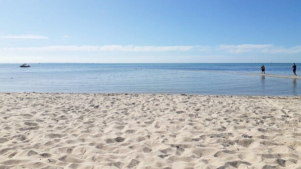 Una playa arenosa con huellas que se dirigen al agua, un barco a lo lejos y un cielo despejado.