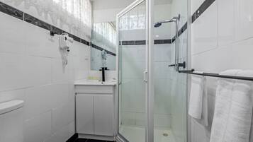 Un baño moderno con ducha acristalada, azulejos blancos, lavamanos con artículos de aseo y un perchero para toallas.