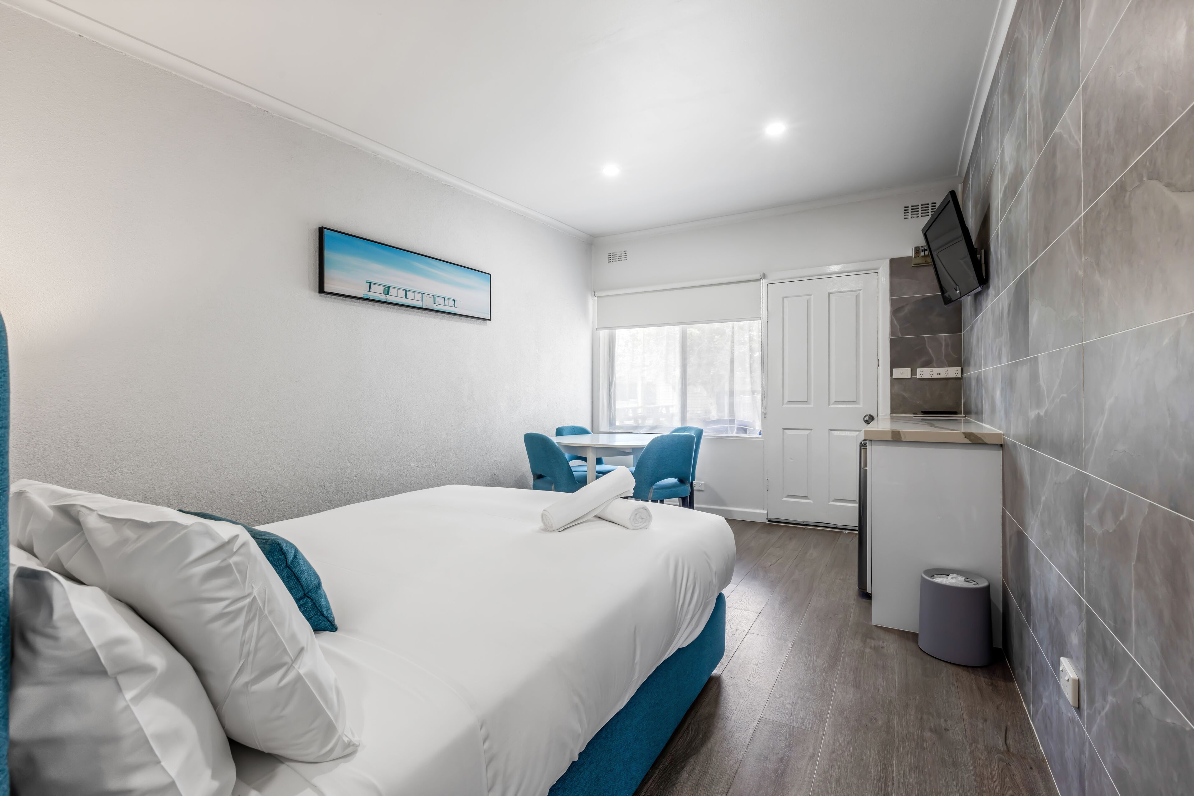 A hotel room with a bed, a desk, a chair, a small kitchen area, and a window.