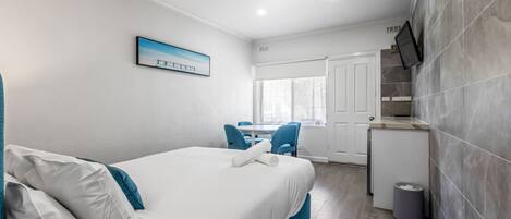 A hotel room with a bed, a desk, a chair, a small kitchen area, and a window.