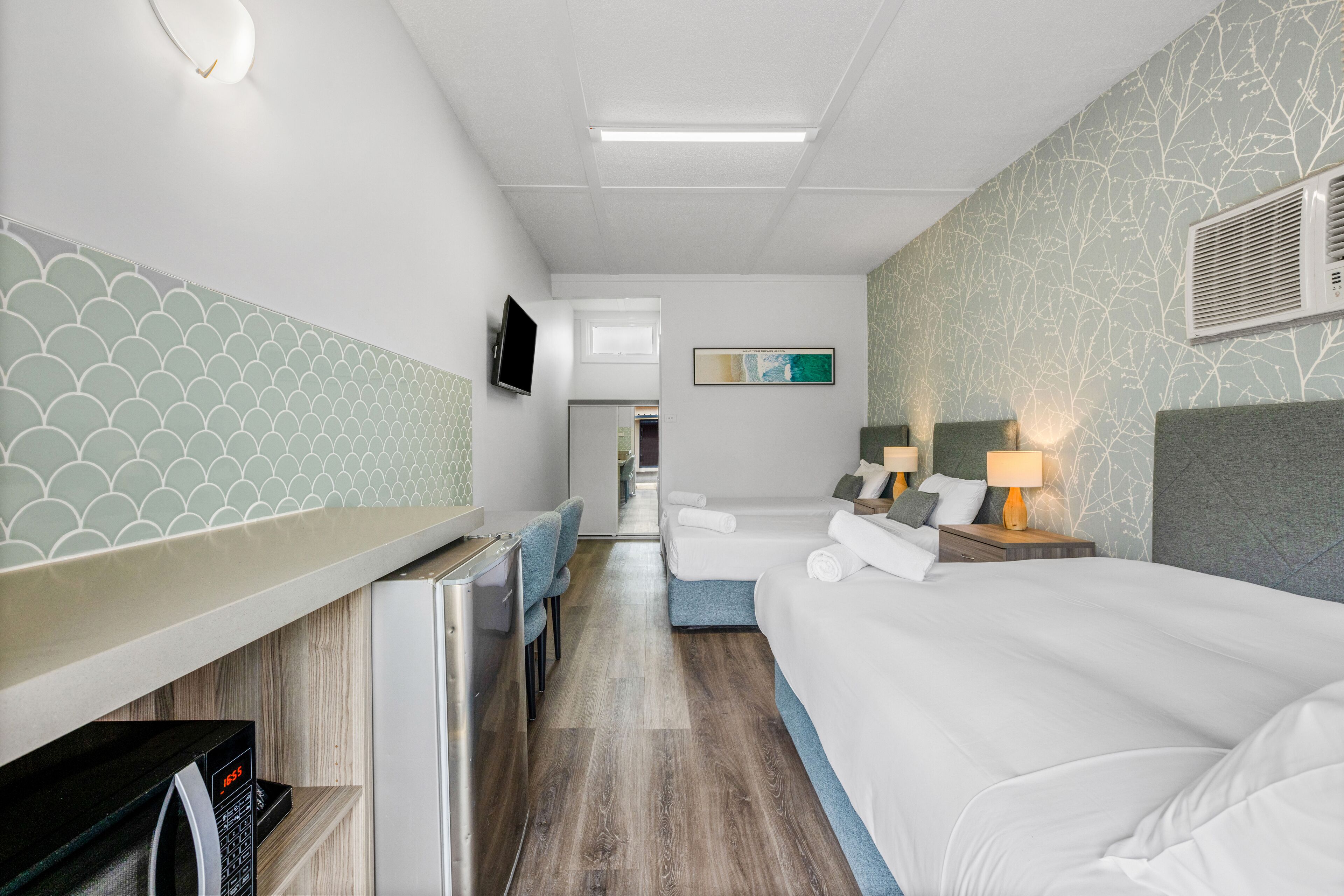 A hotel room with a kitchenette, a TV, a bed with white linens, a desk with a chair, and a bathroom visible through an open door.