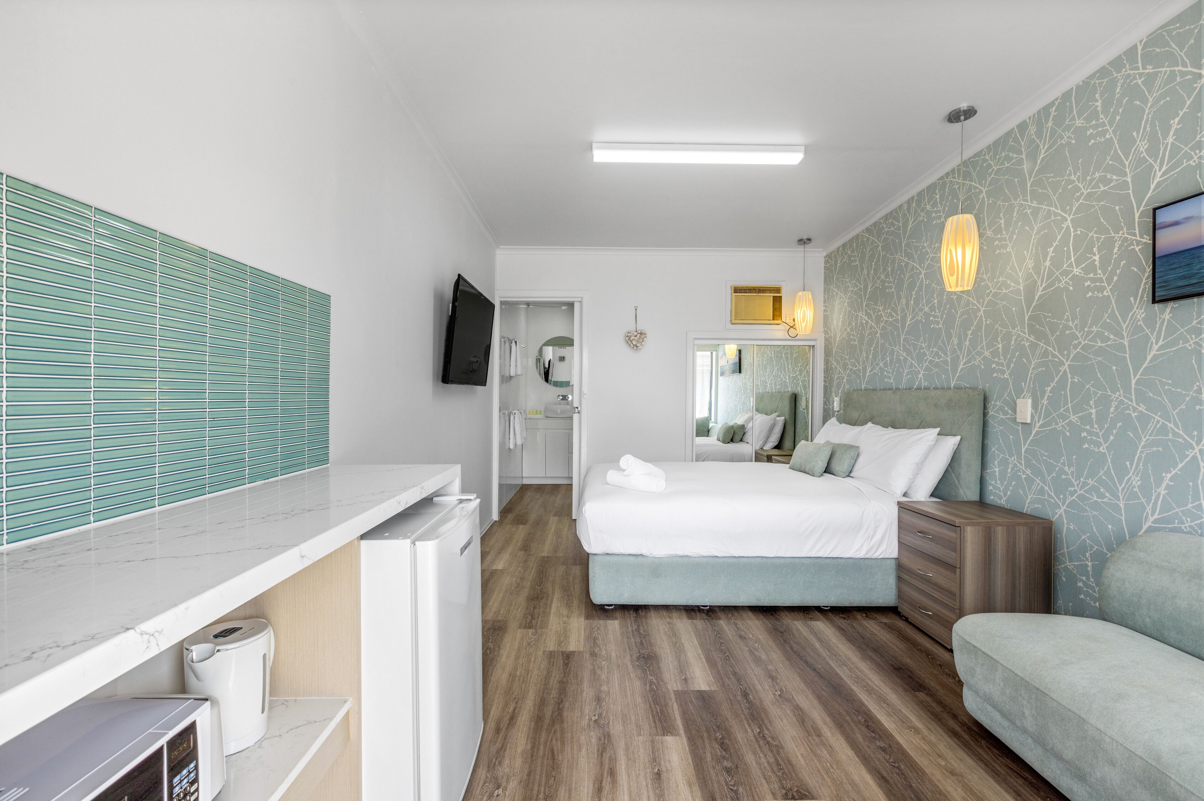 Et moderne hotelværelse med seng, sofa, fjernsyn, spejl og et badeværelse, der kan ses igennem en åben dør.