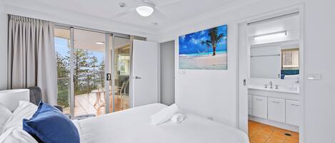 Una habitación de hotel con cama, ventilador de techo, pintura de playa, baño con lavamanos y espejo, y puerta corrediza de vidrio a un espacio exterior.