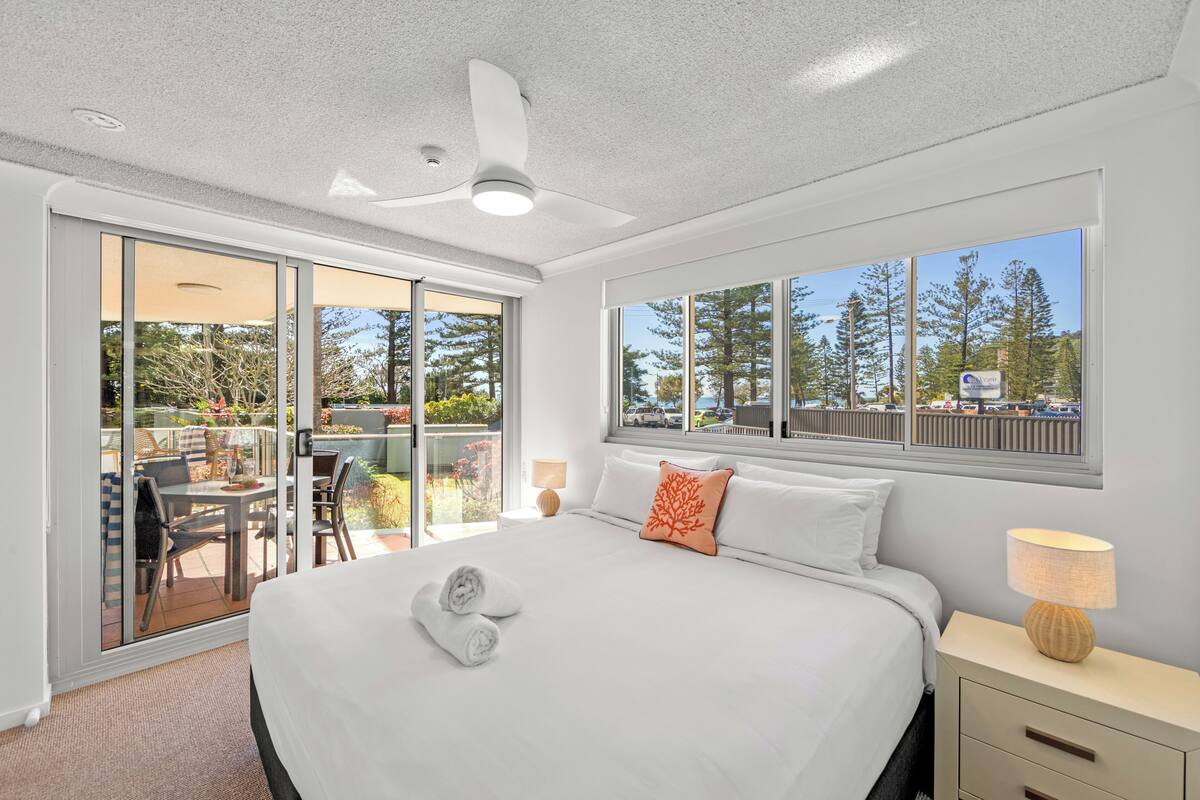 A bedroom with a large bed, a ceiling fan, a nightstand with a lamp, and a view of the outdoors through a sliding glass door.