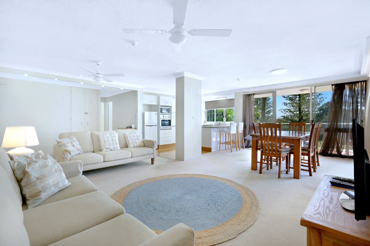 A spacious living area with a sofa, a coffee table, and a dining table with chairs. There is a ceiling fan, a kitchen in the background, and large windows with curtains.