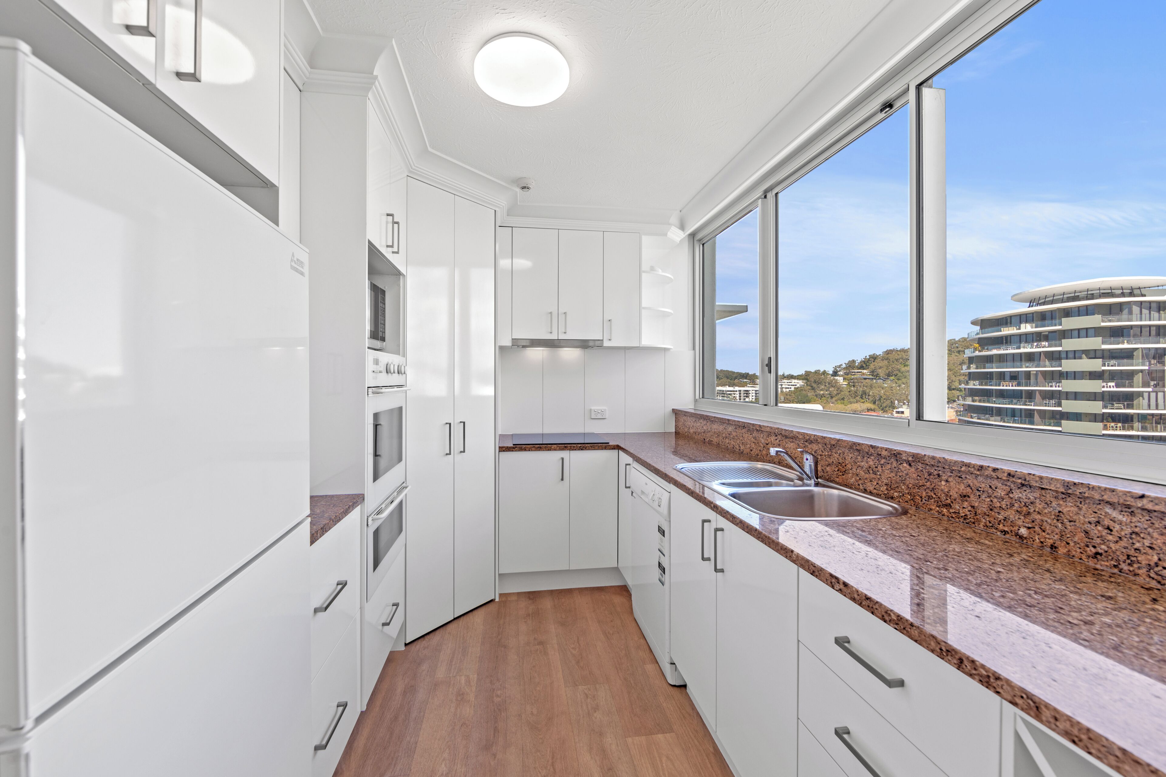 Una cocina moderna con armarios blancos, encimera de granito y un amplio ventanal con vistas a un edificio.