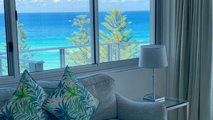A modern living room with a grey sofa, tropical patterned cushions, a glass coffee table, and a view of the ocean through large windows. - Pacific Regis Apartments (Burleigh Heads)