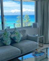 Una sala moderna con un sofá gris, cojines estampados de motivos tropicales, una mesa de centro de vidrio y vistas al océano a través de grandes ventanales.