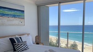 A hotel room with a large bed, a ceiling fan, a beach view through the window, and a painting on the wall. - Pacific Regis Apartments (Burleigh Heads)