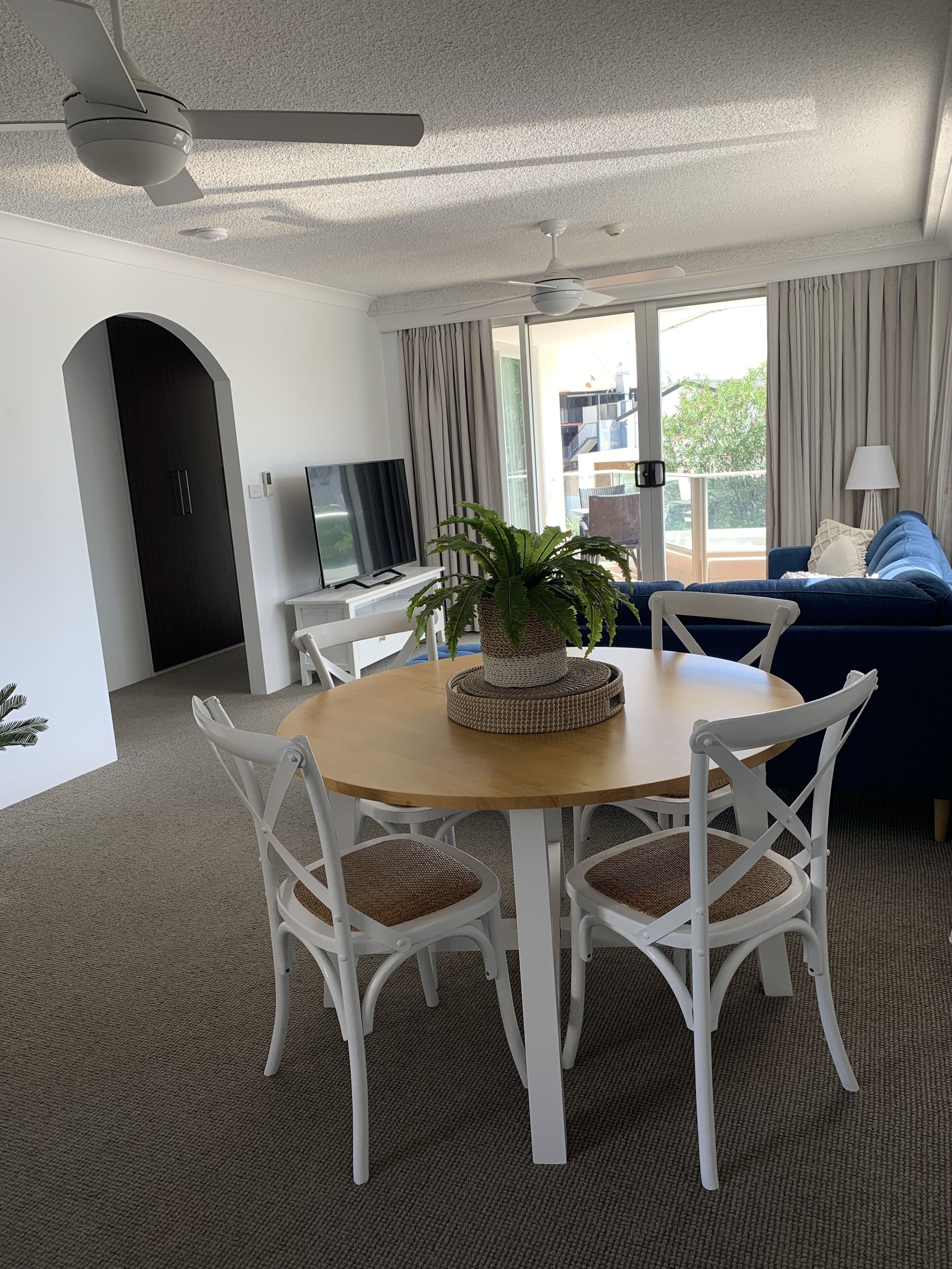 A modern living room with a round wooden table, white chairs, a dark blue sofa, and a TV on a white stand.