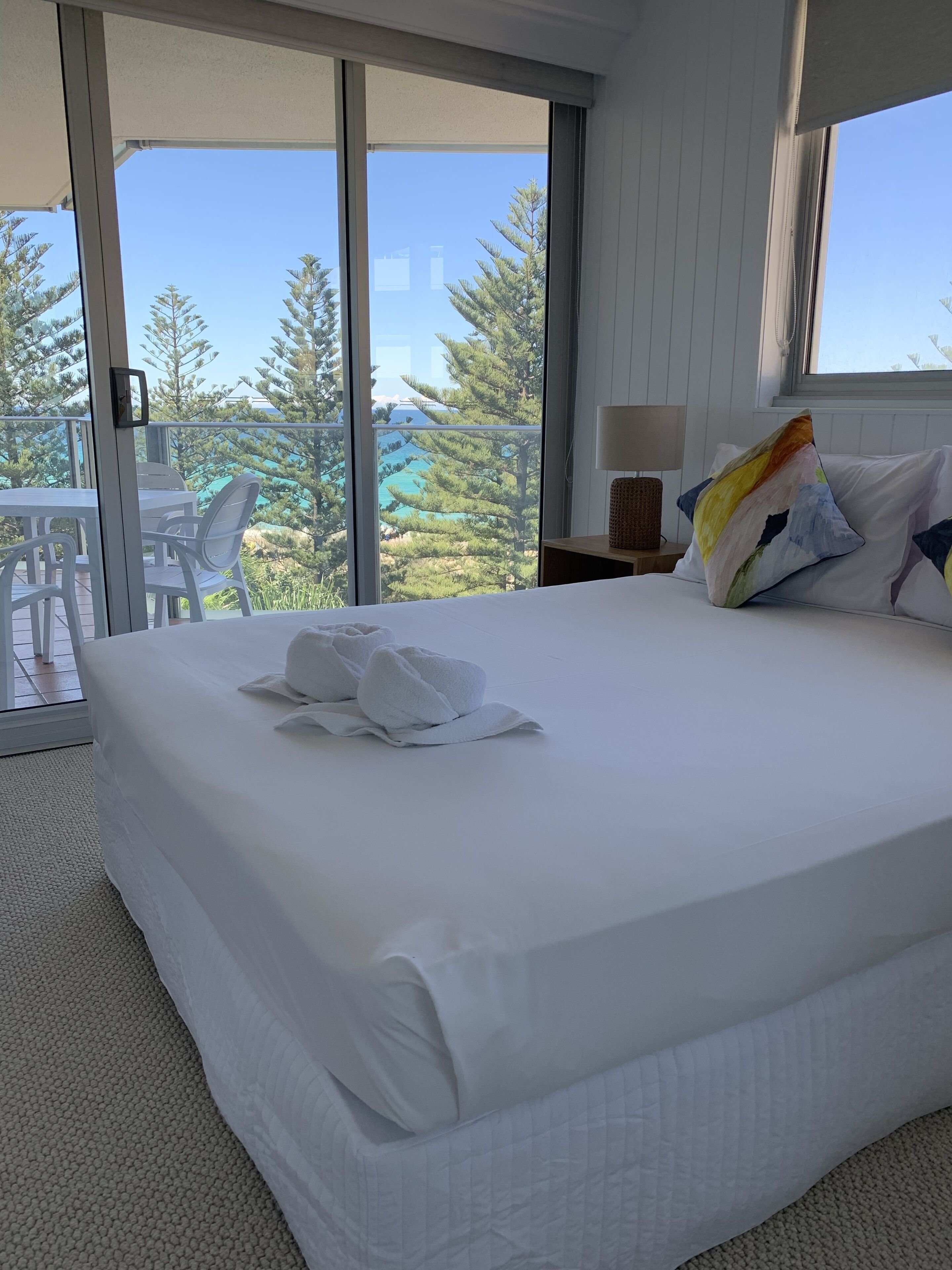 Hotelkamer met een groot bed, wit beddengoed, uitzicht op bomen en een zwembad door de balkondeuren, en een kleine tafel met stoelen buiten.