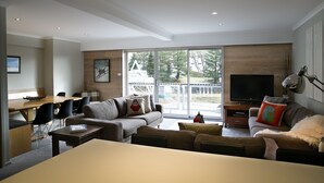 A modern living room with a sofa, coffee table, and TV. There is a dining area with chairs and a table. The room has large windows and a view of the outside. - Altitude Apartments (Falls Creek)
