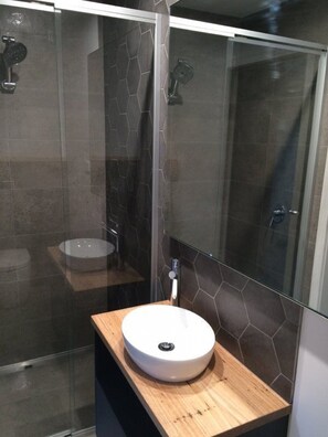 Un baño moderno con ducha envuelta en vidrio, un lavamanos redondo sobre una encimera de madera y azulejos hexagonales en las paredes.