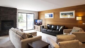 A modern living room with a stone fireplace, leather sofas, a flat-screen TV, and wooden paneling. - Altitude Apartments (Falls Creek)