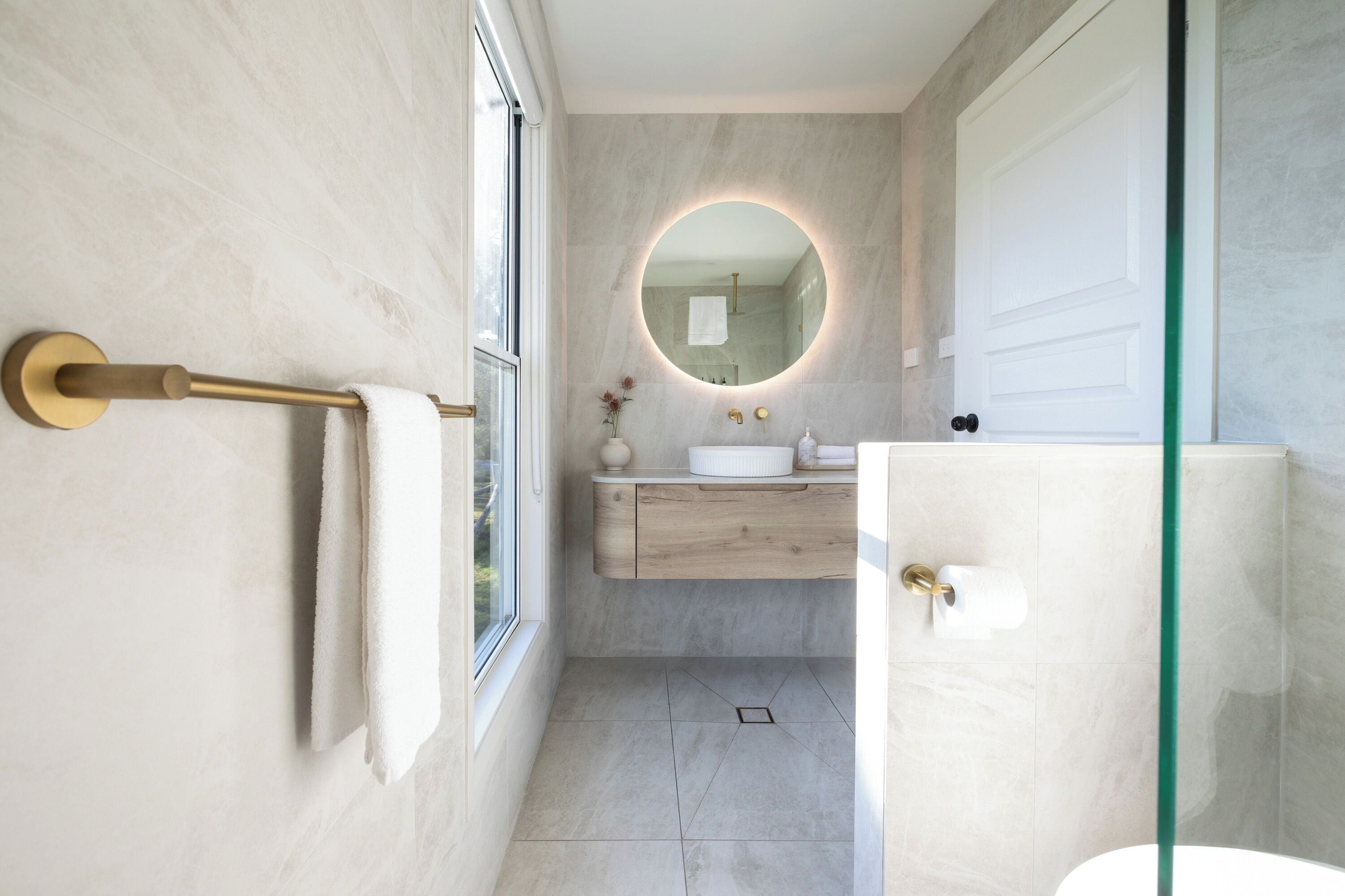 A modern bathroom with a marble wall, a round mirror, a wooden vanity, and a glass shower enclosure.