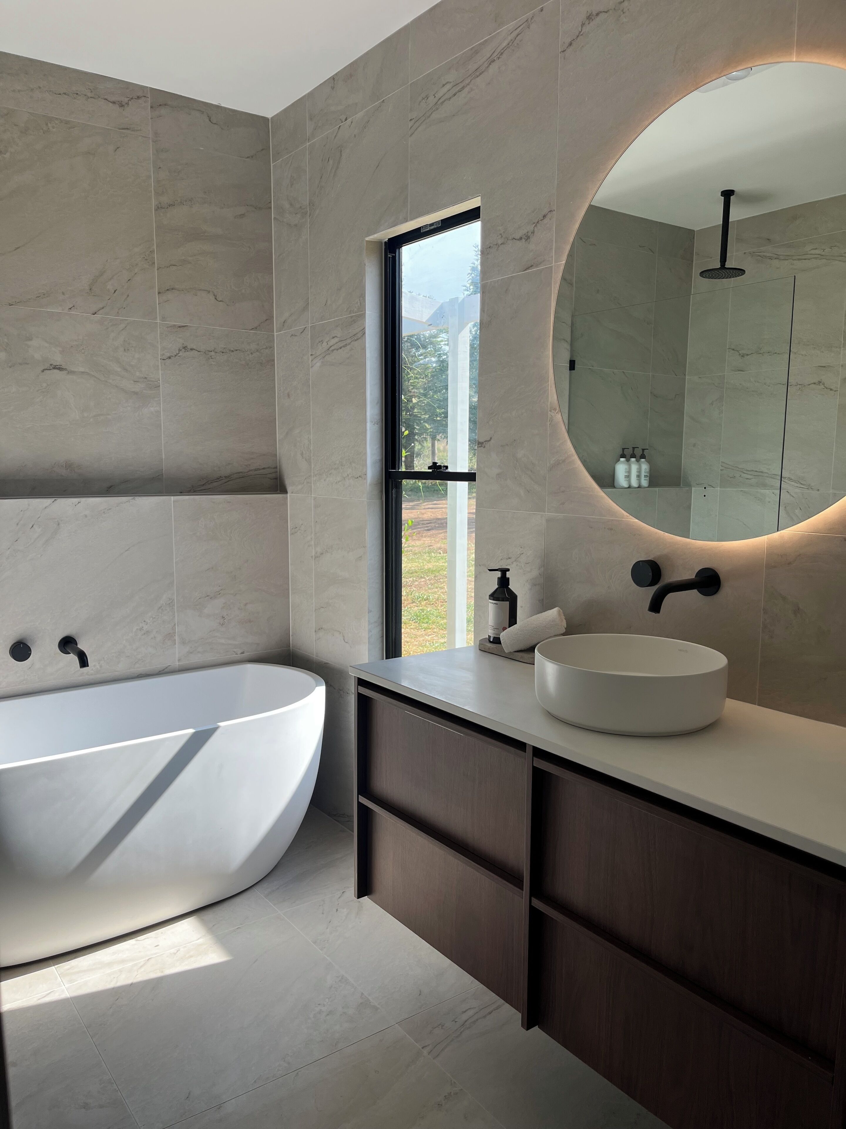 Un bagno moderno con una vasca freestanding, uno specchio tondo con cornice illuminata e una finestra che offre vista sull'esterno.