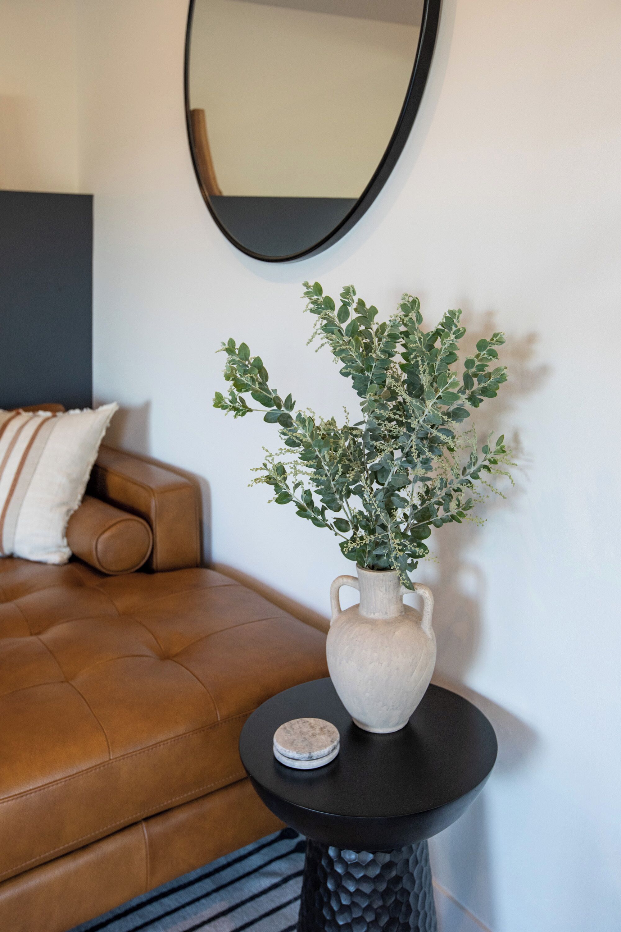 Ein braunes Ledersofa mit einem weißen Kissen, ein kleiner runder Tisch mit einer Vase mit Grünpflanzen und ein runder Spiegel an der Wand.