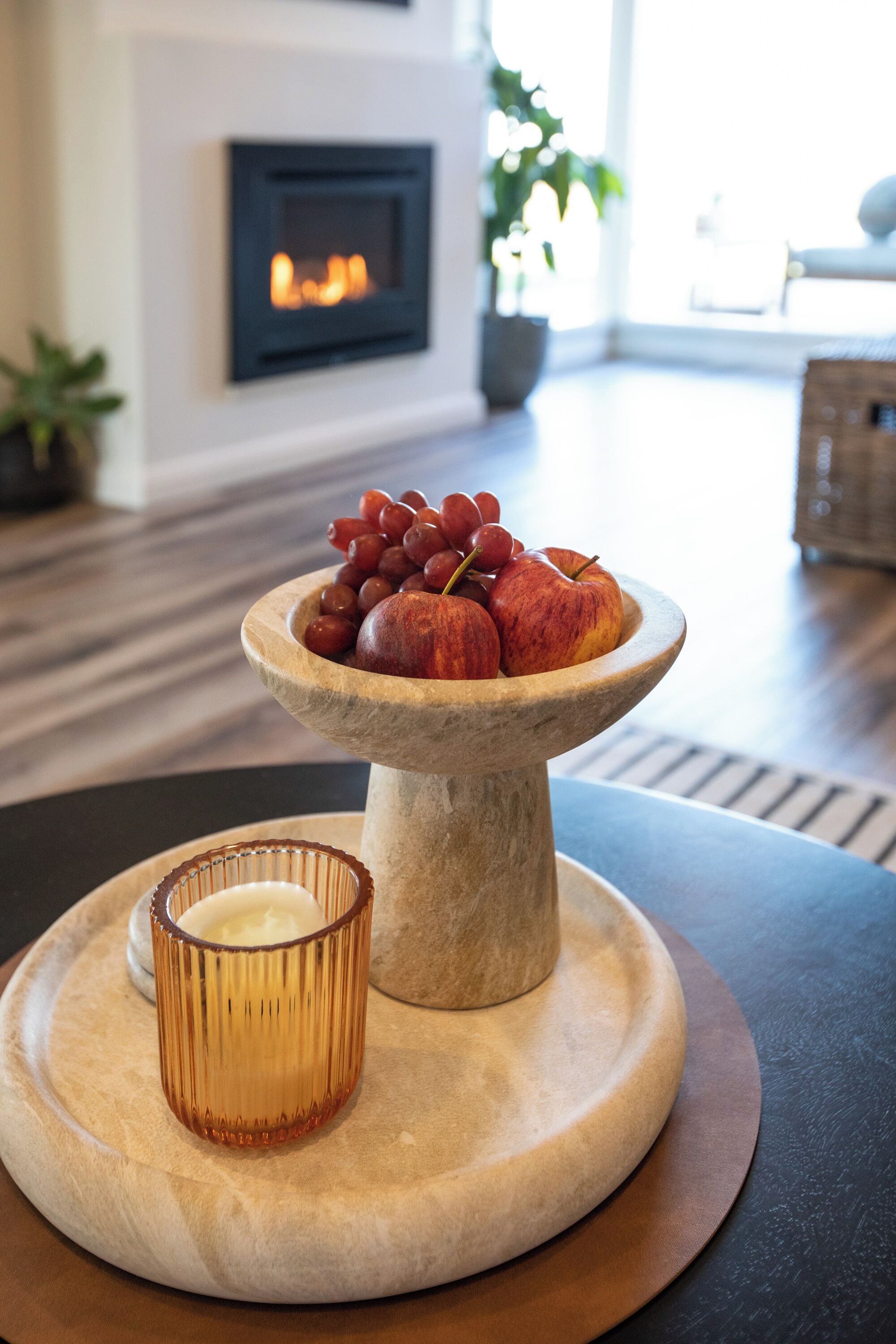 Une table à manger avec un panier de fruits contenant des raisins et des pommes, une bougie et une cheminée en arrière-plan.