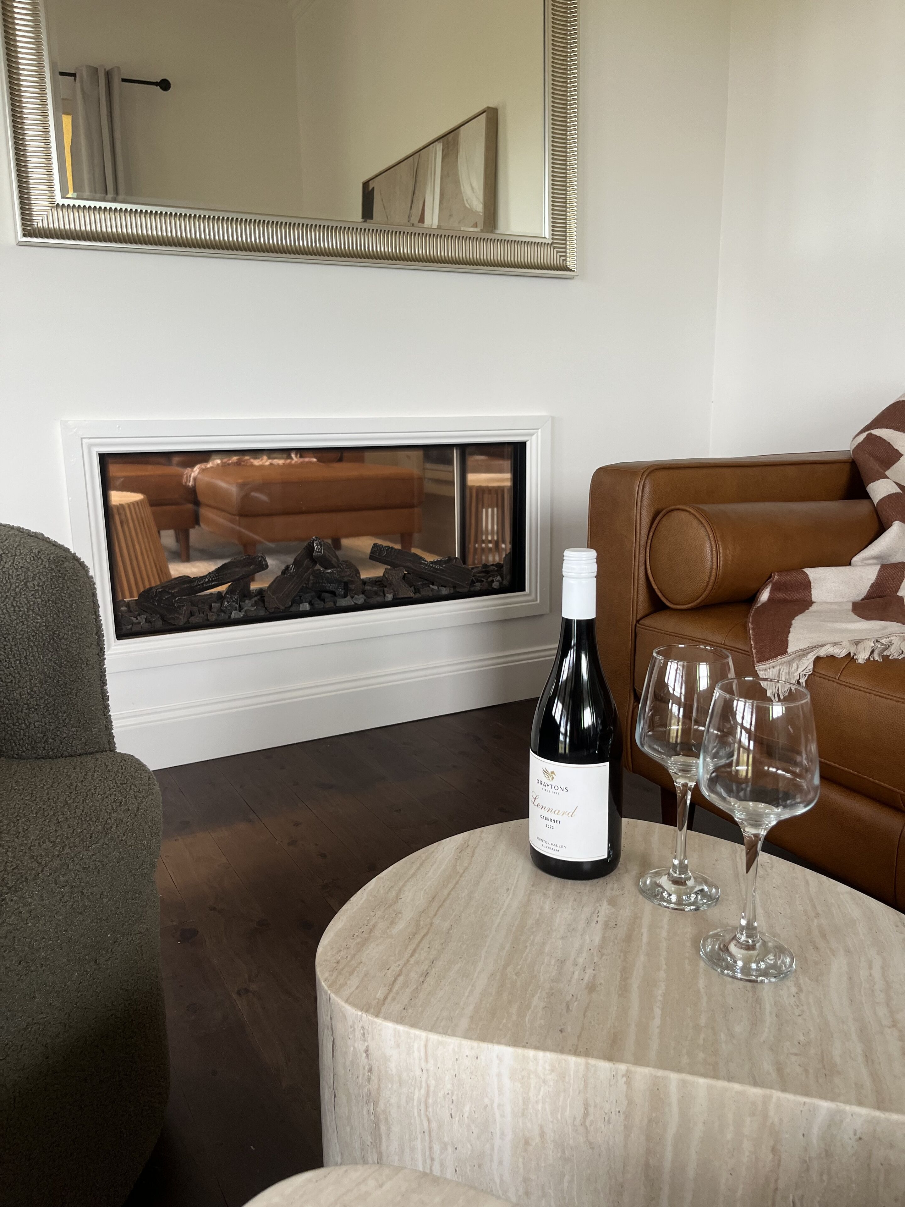 A living room with a fireplace, a brown leather sofa, a round marble table with a wine bottle and glasses, and a large mirror on the wall.